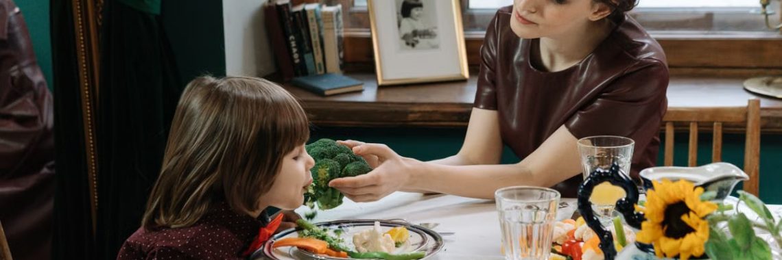 A caring mother helps her child eat healthy vegetables at a dining table, promoting family bonding.